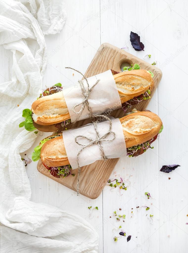 Sandwiches with beef, fresh vegetables and herbs on rustic wooden chopping board over white backdrop