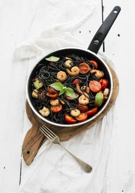 Black pasta spaghetti with shrimps, basil, pesto sauce and slow-roasted cherry-tomatoes in cooking pan on rustic chopping board over white wooden table clipart