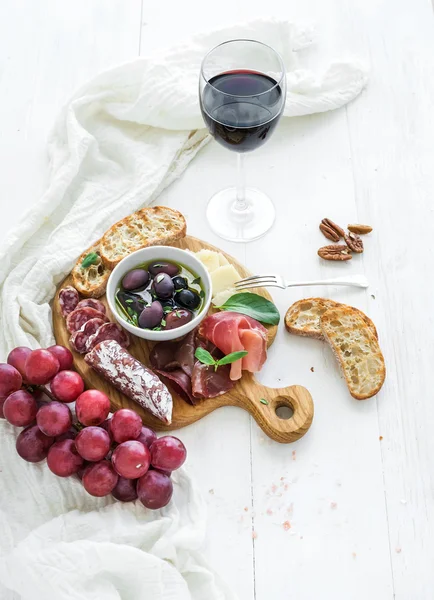 Wine appetizer set. Glass of red, grapes, parmesan cheese, meat variety, bread slices, pecan nuts, honey, olives and basil on rustic wooden board over white backdrop