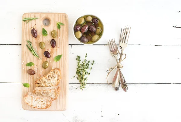 Aceitunas mediterráneas con hierbas y rodajas de ciabatta sobre tabla de madera rústica sobre fondo blanco, vista superior —  Fotos de Stock