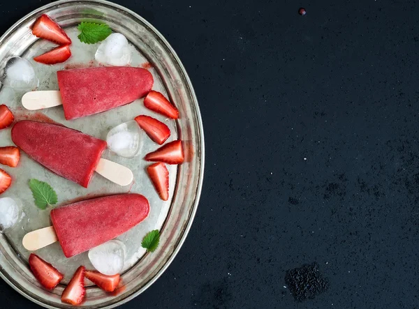 Strawberry ice-creams or popsicles with fresh berries, ice cubes and melissa leaves on vintage silver tray over dark grunge backdrop, top view