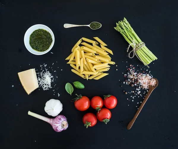 Ingredients for cooking pasta. Penne, green asparagus, basil, pesto sauce, garlic, spices, parmesan cheese and  cherry-tomatoes on dark grunge backdrop — Stock Photo, Image