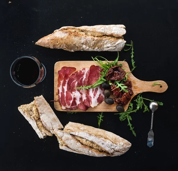 Wine appetizer set: vintage dinnerware, french baguette broken into pieces, dried tomatoes, olives, smoked meat and arugula on rustic wooden board over dark background — Stock Photo, Image