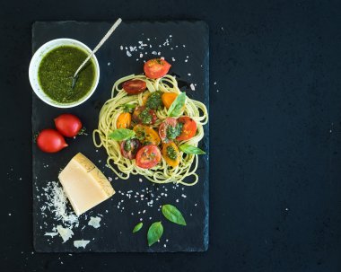 Spaghetti with pesto sauce, roasted cherry-tomatoes, fresh basil and parmesan cheese on black stone serving board over dark grunge backdrop. Top view