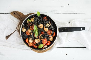 Black pasta spaghetti with shrimps, basil, pesto sauce and slow-roasted cherry-tomatoes in cooking pan on rustic chopping board over white wooden table clipart