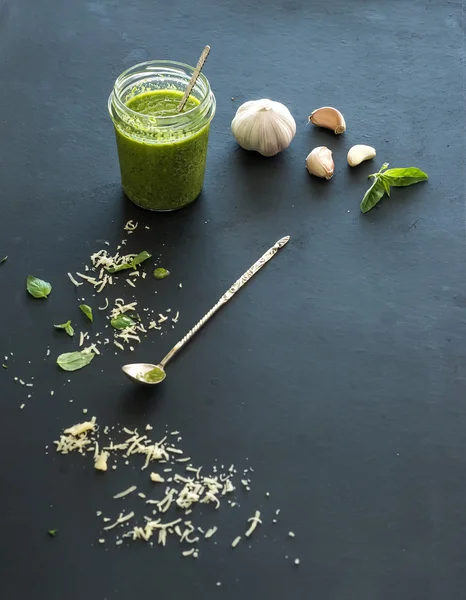 Frasco de pesto caseiro fresco com ingredientes para ele. Folhas frescas de manjericão, queijo parmesão ralado e alho sobre fundo preto. Vista superior — Fotografia de Stock