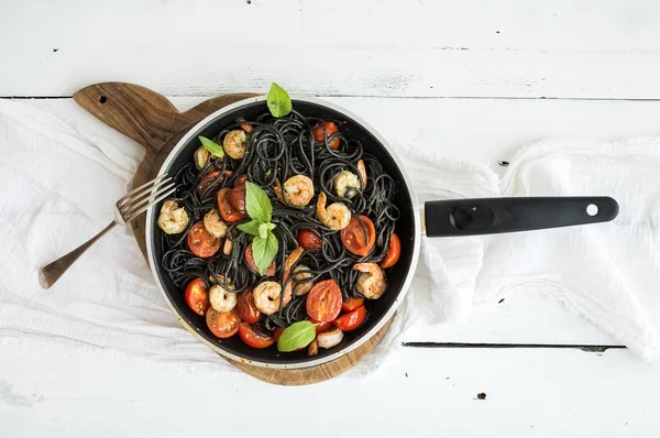 Чорна паста спагетті з креветками, базиліком, соусом песто і повільно обсмаженими вишневими помідорами на сковороді на сільській дошці на білому дерев'яному столі — стокове фото