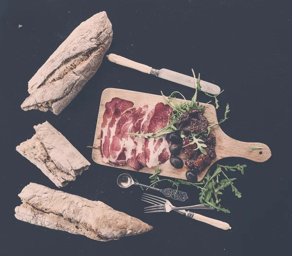 Set de aperitivos de vino: vajilla vintage, baguette francesa desmenuzada, tomates secos, aceitunas, carne ahumada y rúcula sobre tabla de madera rústica sobre fondo oscuro — Foto de Stock