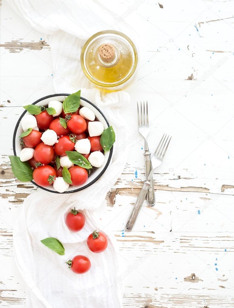 Caprese salad: cherry-tomatoes and mozzarella in metal bowl with olive oil on rustic white wooden backdrop, top view,