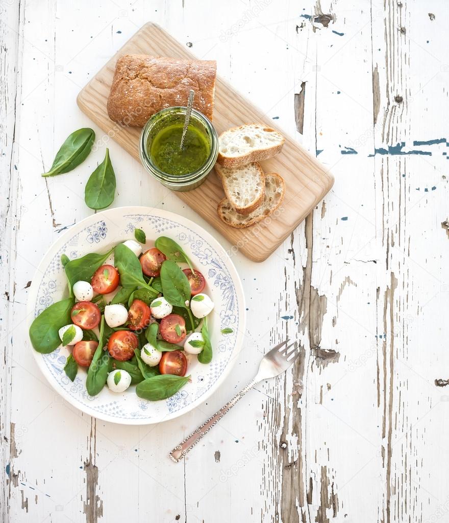 Caprese salad, chiabatta slices, homemade pesto sauce. Cherry-tomatoes, baby spinach and mozzarella in ceramic plate on rustic white wooden backdrop, top view