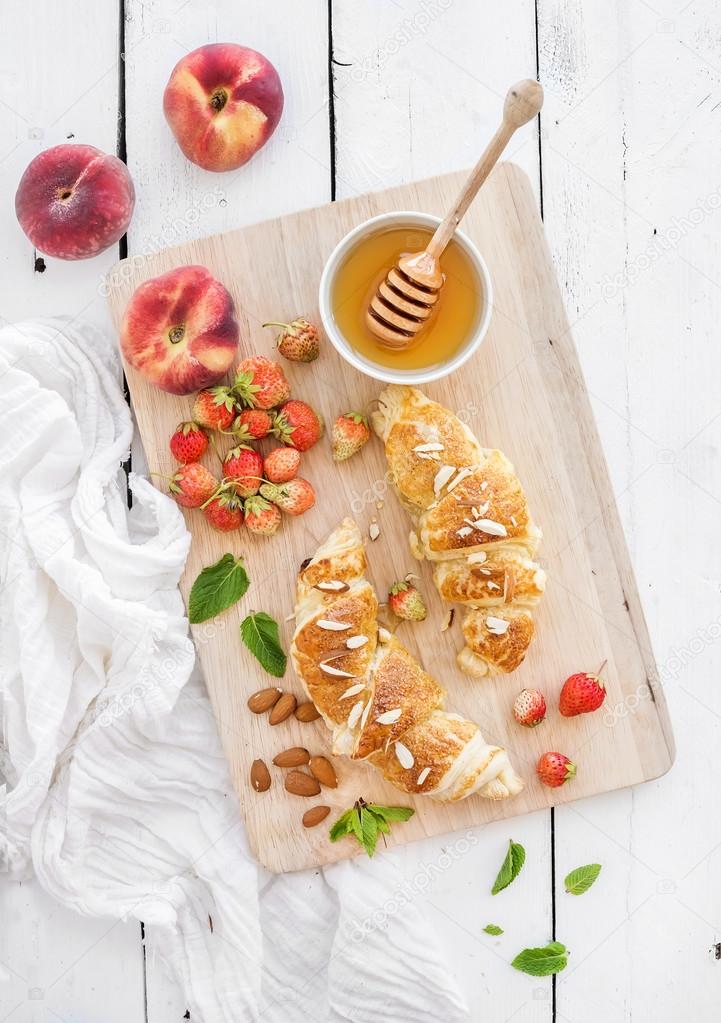 Freshly baked almond croissants with garden strawberries, peaches, mint and honey on serving board over white rustic wood backdrop