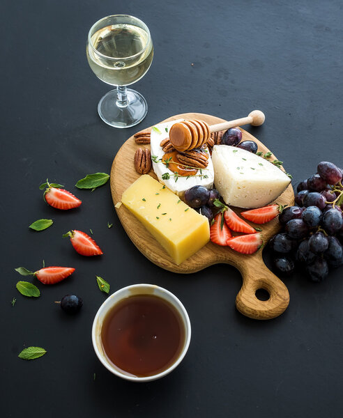 Cheese appetizer selection or whine snack set. Variety of cheese, grapes, pecan nuts, strawberry and honey on round wooden board over black backdrop