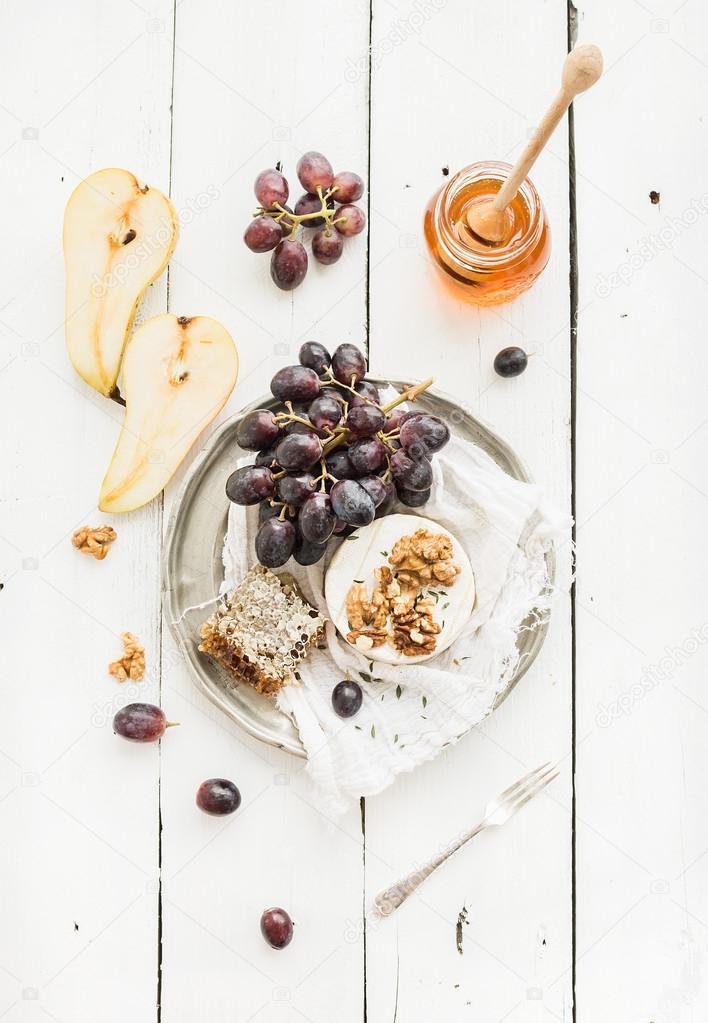 Camembert cheese with grape, walnuts, pear and honey on vintage metal plate over white rustic wood backdrop