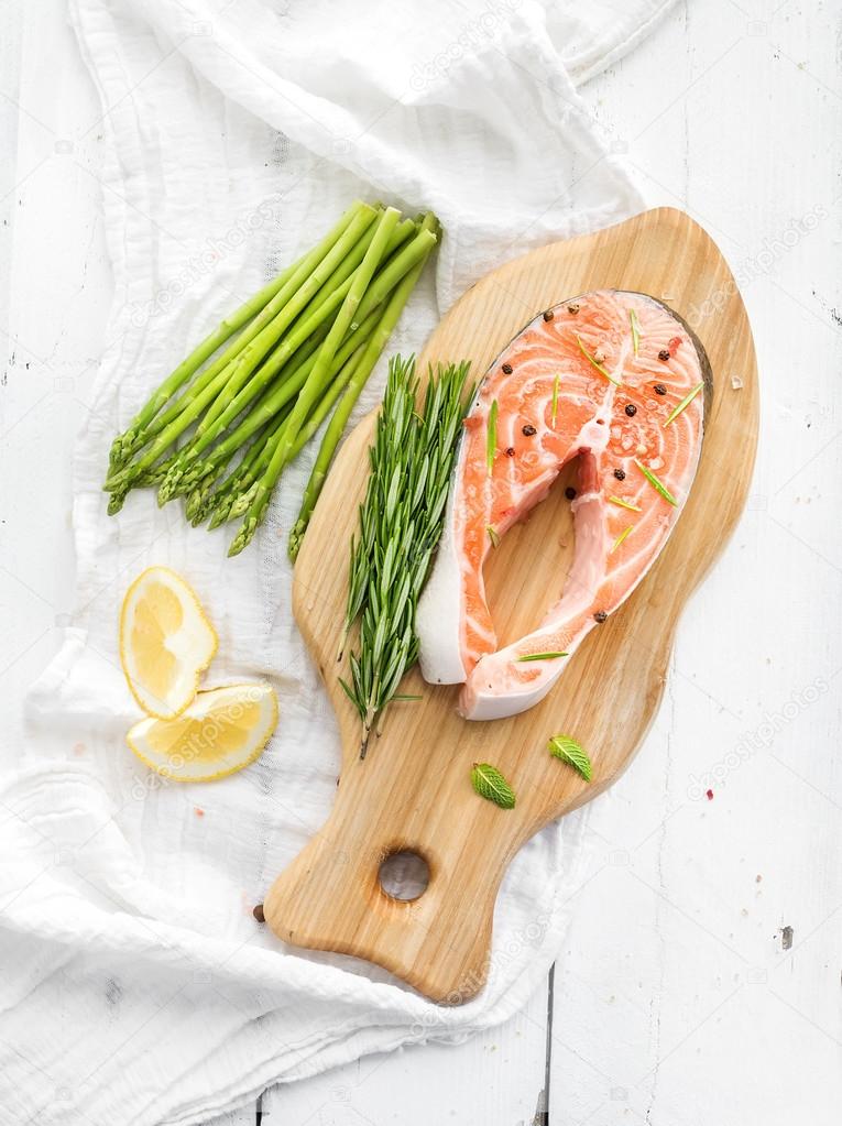 Raw salmon steak with asparagus, lemon, spices and rosemary on rustic wooden chopping board over white backdrop. Top view