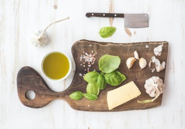 Pesto sauce cooking set. Fresh basil, olive oil, parmesan cheese and garlic on rustic walnut chopping board over white wooden backdrop