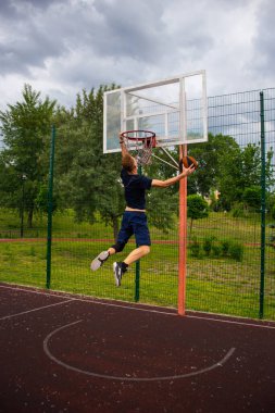 Koyu mavi tişörtlü genç, kızıl saçlı adam topu mavi gökyüzüne doğru basket potasına fırlatıyor.