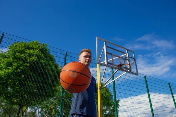 A young red-haired guy in a dark blue t-shirt stands with a basketball ball in his hand on the sports ground with a boxetball ring on a mesh background