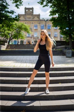 Siyah tişörtlü ve siyah dar spor şortlu güzel sarışın kadın merdivenlerde dikiliyor ve düz görünüyor.