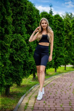 Siyah tişörtlü, siyah dar şortlu, yeşil ağaçların ve çalıların önünde poz veren güzel sarışın bir kadın.