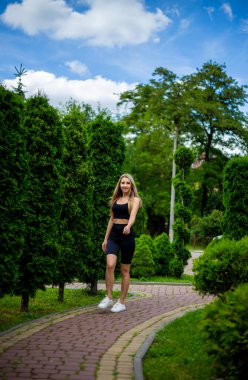 Siyah tişörtlü, siyah dar şortlu, yeşil ağaçların ve çalıların önünde poz veren güzel sarışın bir kadın.