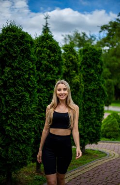 Siyah tişörtlü, siyah dar şortlu, yeşil ağaçların ve çalıların önünde poz veren güzel sarışın bir kadın.