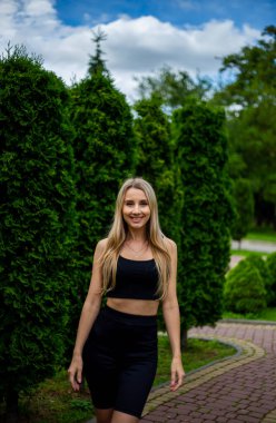 Siyah tişörtlü, siyah dar şortlu, yeşil ağaçların ve çalıların önünde poz veren güzel sarışın bir kadın.