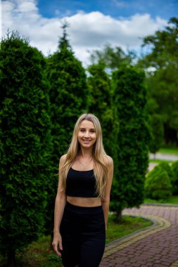 Siyah tişörtlü, siyah dar şortlu, yeşil ağaçların ve çalıların önünde poz veren güzel sarışın bir kadın.