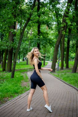 Siyah tişörtlü, siyah dar şortlu, yeşil ağaçların ve çalıların önünde poz veren güzel sarışın bir kadın.