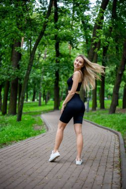 Siyah tişörtlü, siyah dar şortlu, yeşil ağaçların ve çalıların önünde poz veren güzel sarışın bir kadın.