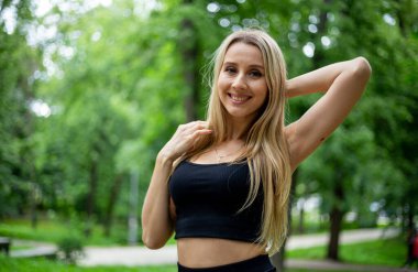 Siyah tişörtlü, siyah dar şortlu, yeşil ağaçların ve çalıların önünde poz veren güzel sarışın bir kadın.