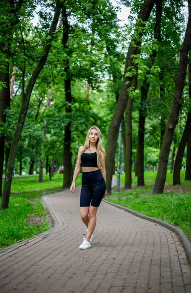 身穿黑色T恤和黑色紧身衣短裤 身材匀称的年轻而漂亮的金发女运动员 站在绿树成荫的大自然面前 — 图库照片