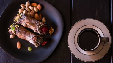 Siyah tabakta iki cannoli, krem peynirden yapılmış geleneksel İtalyan tatlısı ve fıstık ezmeli kahve..