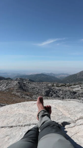 Dağ manzarası, dağın tepesinde dinlenen bir adamın ayakları, arka planda daha fazla dağ zirveleri ve mavi gökyüzü var.