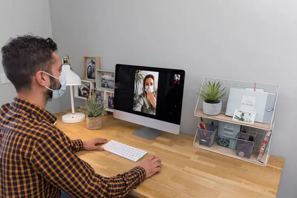 man makes a video call with a woman, dark-haired man in a plaid shirt makes a video call with a woman from the computer, both wear masks