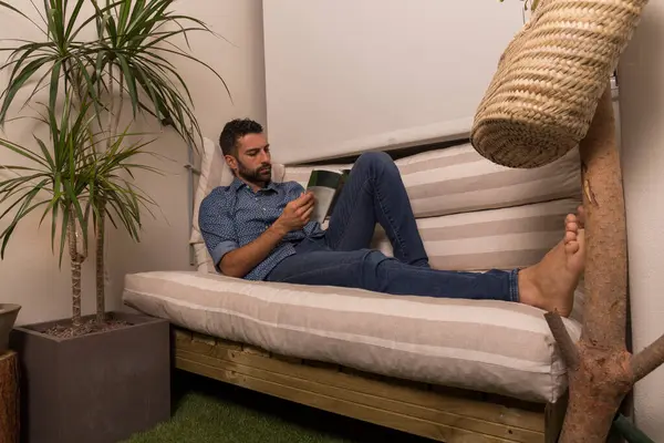 young man rests on a sofa while reading a book, dark man with a beard lying on a striped sofa, rests after a long day at work, next to the sofa there are plants