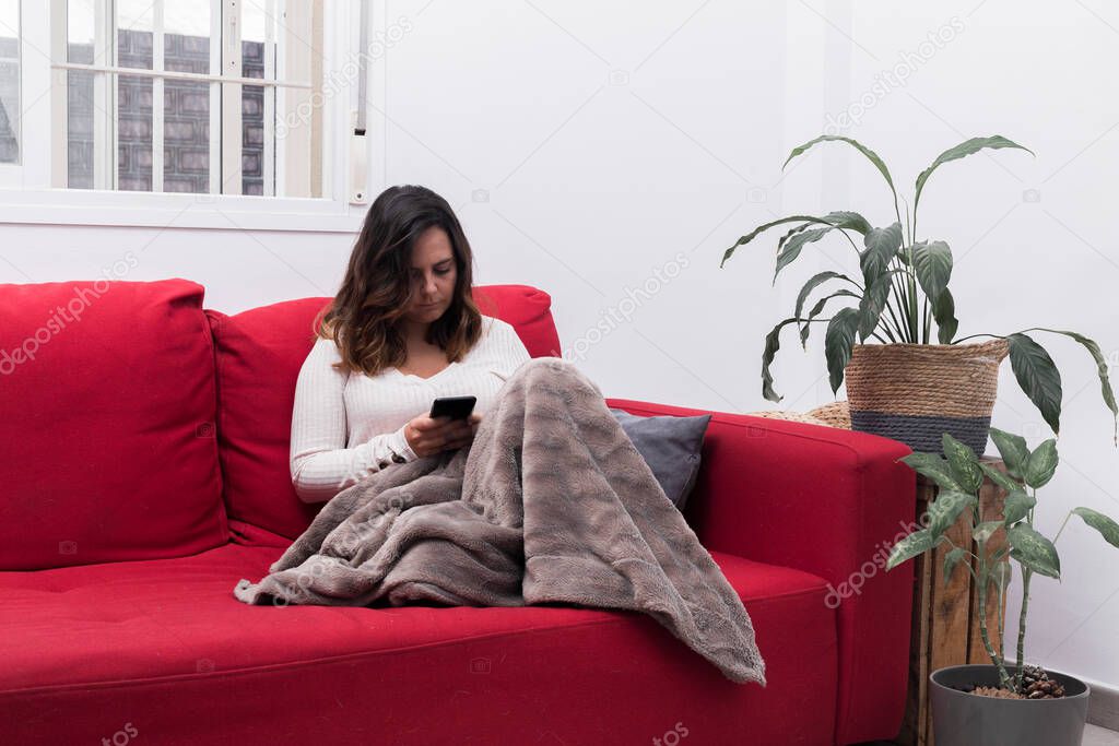 woman sends messages with the mobile, brunette woman sitting on a red sofa and covered with a blanket writes with the mobile, next to the sofa there are plants, horizontal photo