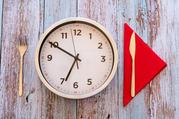 The concept of intermittent fasting and skipping breakfast - large clock with cutlery and red napkin on rustic wooden background. Concept of eight-hour feeding window or breakfast time concept.