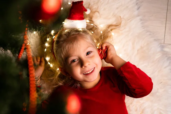 Feliz niño sonriente sosteniendo un regalo bajo el árbol de Navidad en casa. Bebé en un vestido rojo de Navidad — Foto de Stock