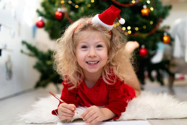 Hermosa niña rubia se encuentra debajo de un árbol en un vestido rojo de Año Nuevo, sonríe — Foto de Stock