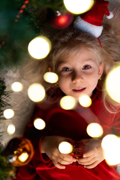 Una hermosa niña rizada yace bajo un árbol de Navidad con luces en el pelo — Foto de Stock