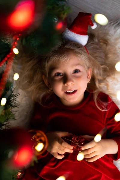 Una hermosa niña rizada yace bajo un árbol de Navidad con luces en el pelo — Foto de Stock