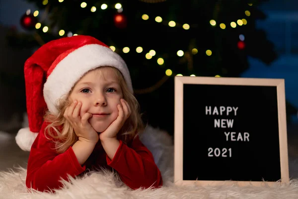 Niña hermosa con sombrero de Santa Claus y vestido de Navidad rojo está acostado bajo el árbol de Navidad — Foto de Stock