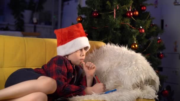 Mały Chłopiec Świątecznym Kapeluszu Pisze List Świętego Mikołaja Leżącego Kanapie — Wideo stockowe