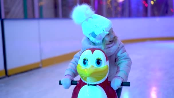 Niña en la pista de hielo, aprendiendo a patinar con el apoyo del simulador de pingüinos y sonriendo — Vídeos de Stock