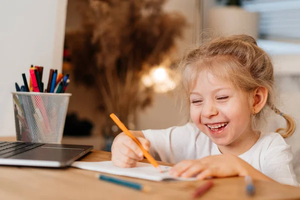 Χαρούμενο κοριτσάκι γελάει με το laptop της και κάνει την εργασία σε ένα σημειωματάριο, σπίτι μάθηση έννοια στην covid 19 πανδημία — Φωτογραφία Αρχείου