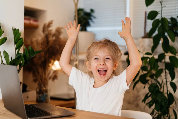 A little girl is sitting at a table with a laptop, raising her hands in the air, smiling and happy, experiencing happiness — Stock Photo, Image