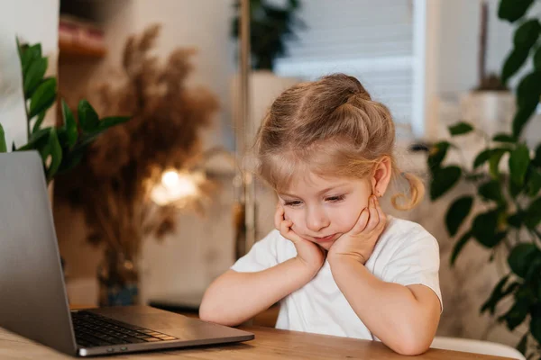 Μικρό σκεπτικό ξανθό κορίτσι κάθεται μπροστά από ένα φορητό υπολογιστή με ένα λυπημένο πρόσωπο, η έννοια της κατ 'οίκον εκπαίδευσης και της μοναξιάς — Φωτογραφία Αρχείου