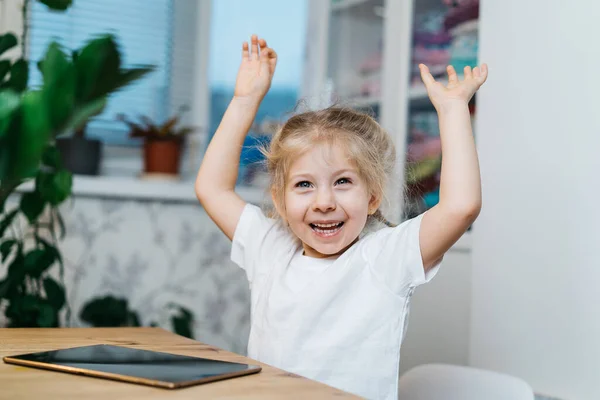Ένα μικρό κορίτσι κάθεται σε ένα τραπέζι με ένα τάμπλετ με τα χέρια ψηλά στον αέρα χαμογελώντας και ευτυχισμένα, βιώνοντας την ευτυχία — Φωτογραφία Αρχείου
