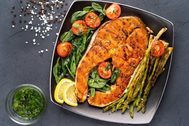 ızgara somon bifteği, kuşkonmaz ve siyah arka planda siyah bir tabakta domatesli taze ıspanak.