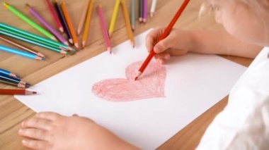 Küçük bir çocuğun elleri beyaz bir kağıt parçasına kırmızı kalemle bir kalp çizer.